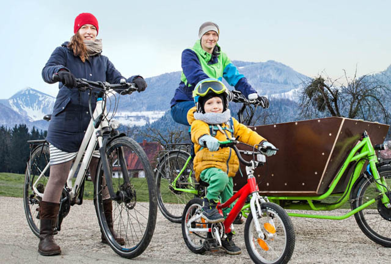 Slide: Pärchen mit Kleinkind fahren mit Lastenrad und Rad vor verschneiten Bergen