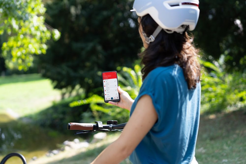 Frau steht vor Fahrrad und schaut aufs Handy darauf sind die mit der Österreich radelt App getrackten Strecken sichtbar. Im Hintergrund Natur