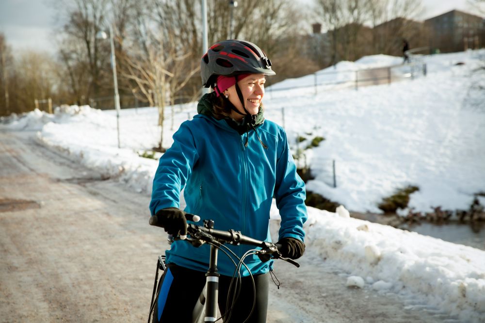 Frau steht mit Rad in Winterkleidung auf Straße und lacht, die Sonne scheint ihr ins Gesicht, Schnee im Hintergrund
